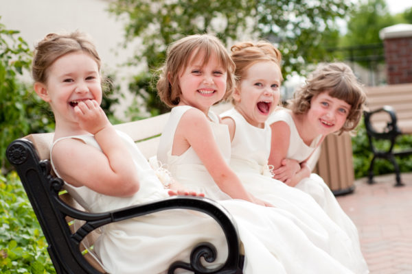 Color photo of four happy little flower girls laughing together while wearing formal dresses. They are sisters and cousins.